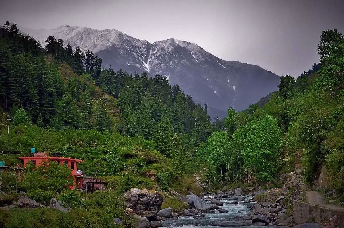 A red-painted house standing above a stony river in dense deodar forest, with a snow-streaked ridge rising behind it under a grey sky.
