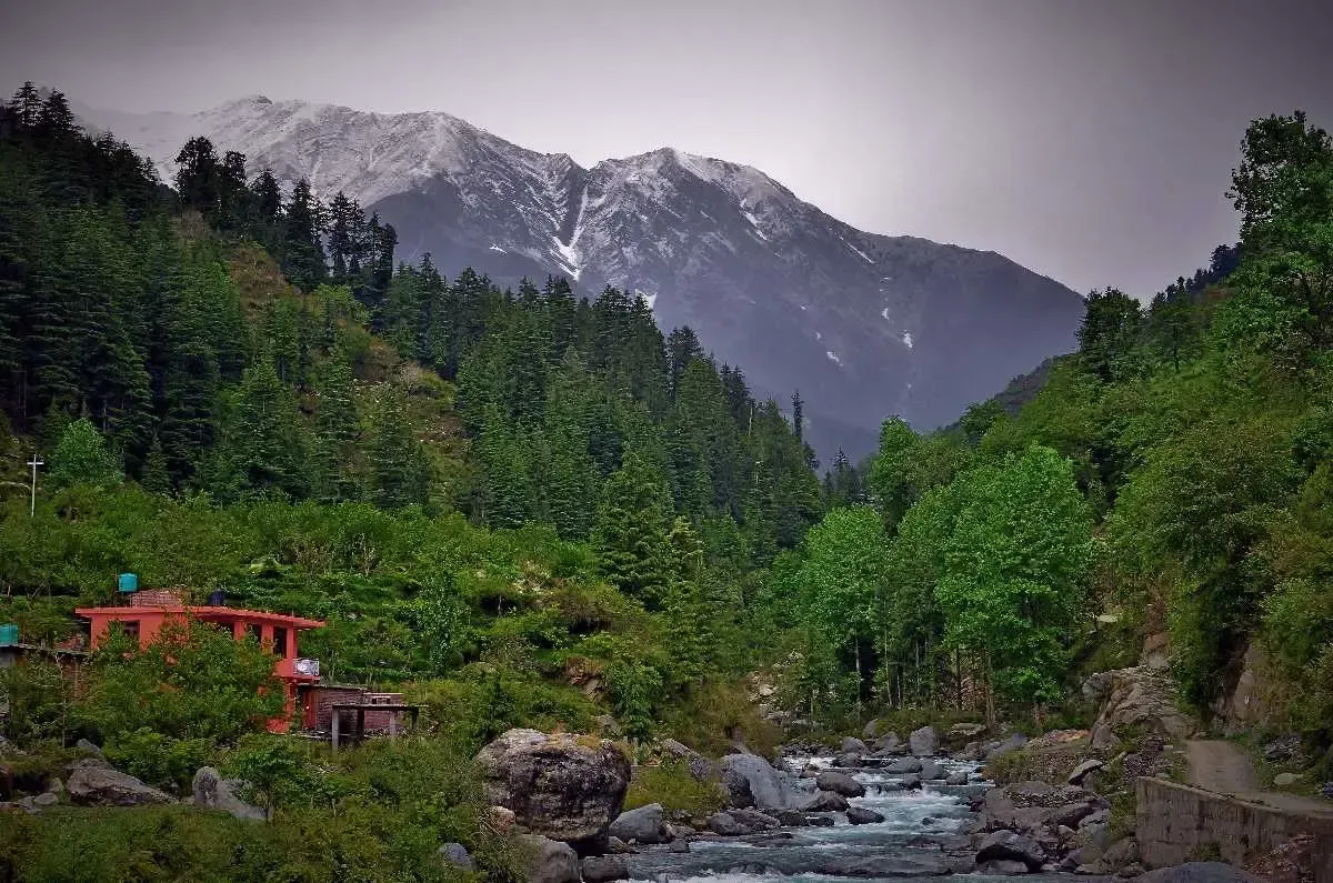 Snow-covered peaks above a steep forested valley, a stream running through boulders below and a red-roofed building among the trees