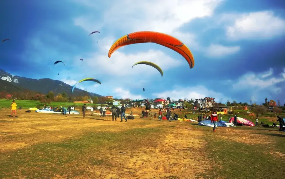 An orange canopy coming in low over a green landing field, spectators and packed gliders spread across the grass