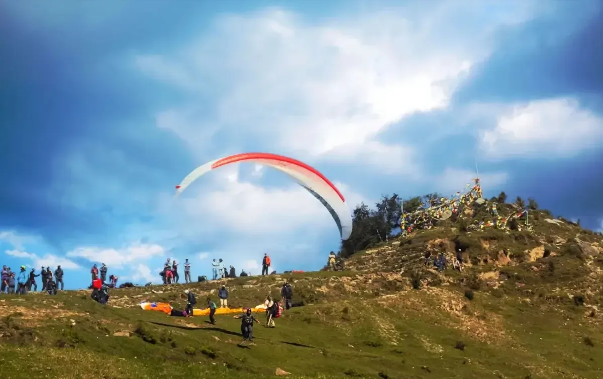 A paraglider lifting off a rocky launch site, a line of people watching from the slope beside it