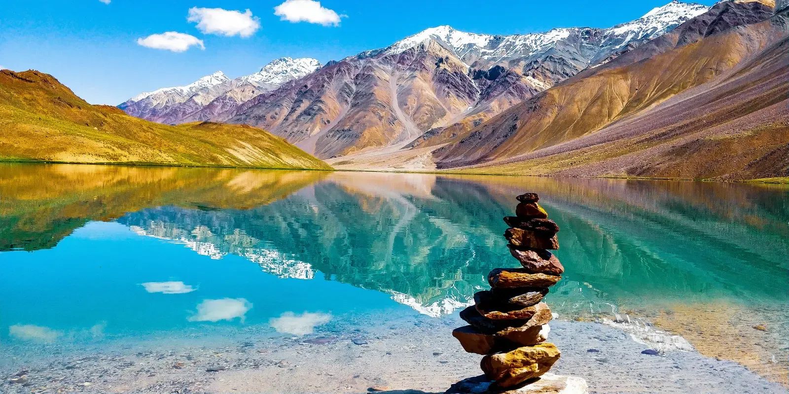 A turquoise lake lying still under bare ochre and purple mountains, the ridge reflected across it and a stack of flat stones balanced on the shore.