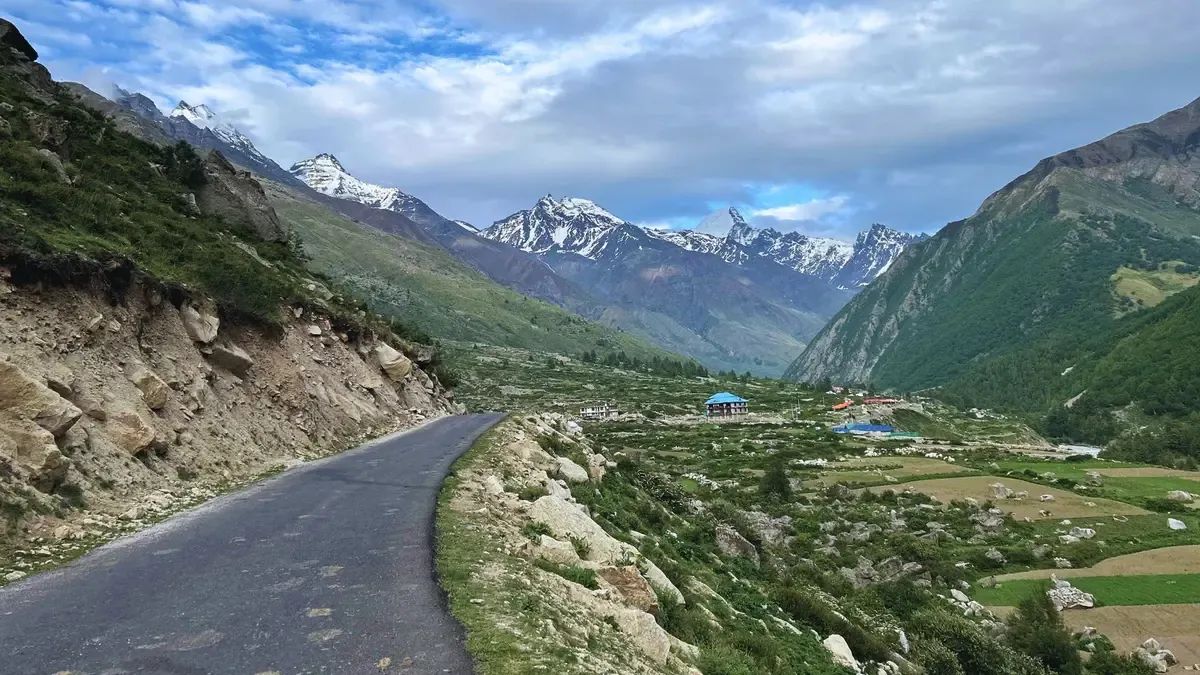 A tarred road curving down a broad green valley between grassy slopes, a cluster of blue and red roofed buildings on the floor of it and snow peaks standing at the head of the valley.