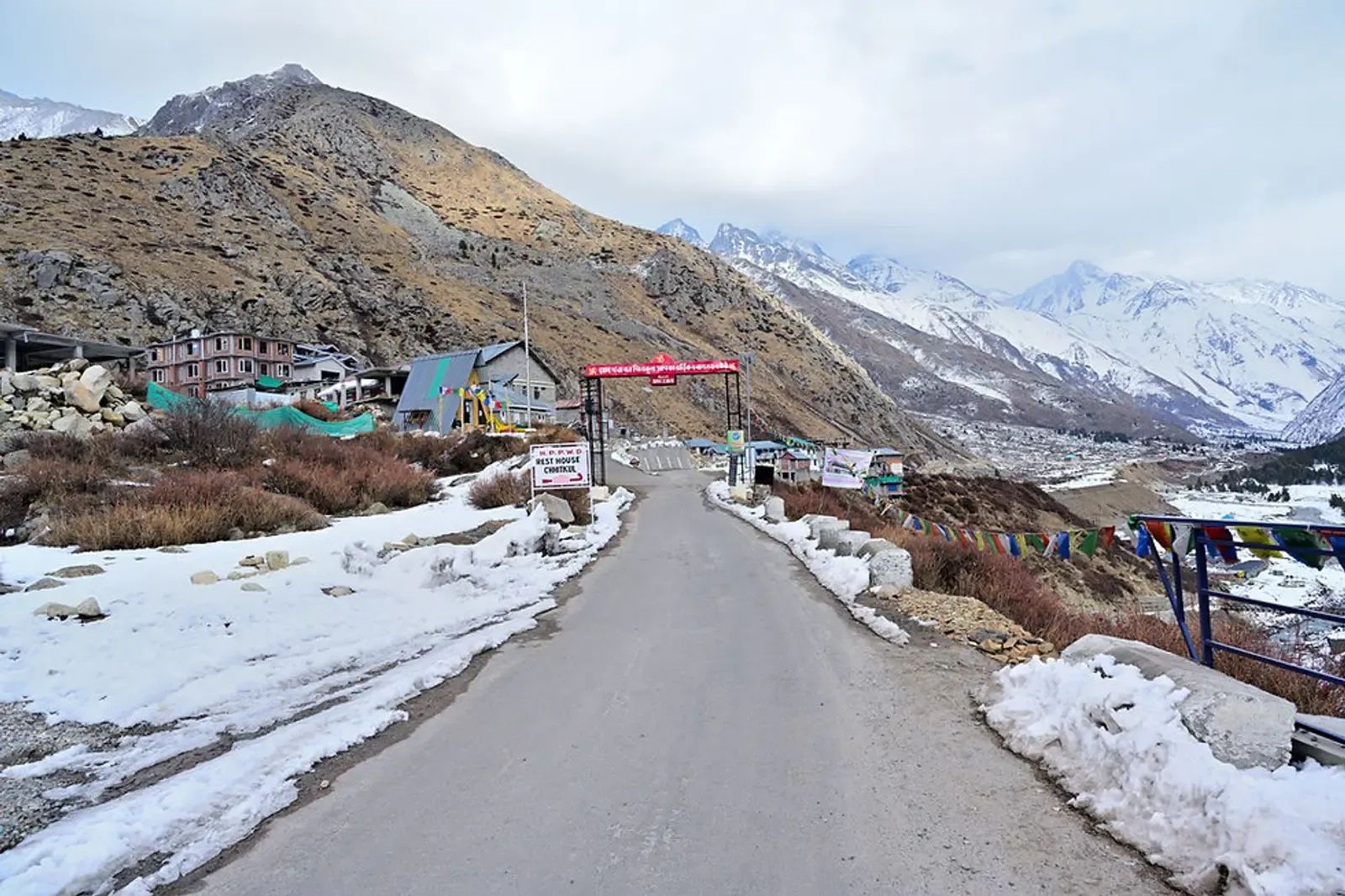 A cleared road between banks of old snow running towards a painted gateway with grey peaks standing behind it.