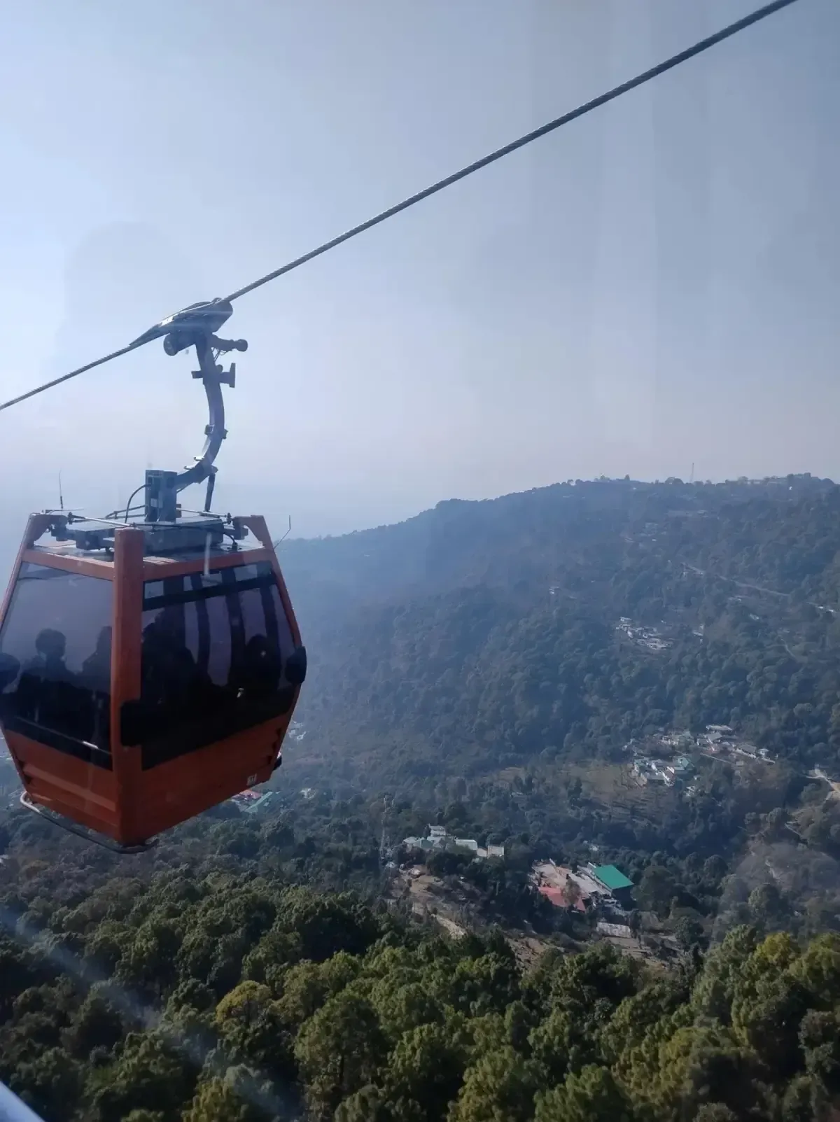 A cable car on a wire above forested slopes with cloud behind it