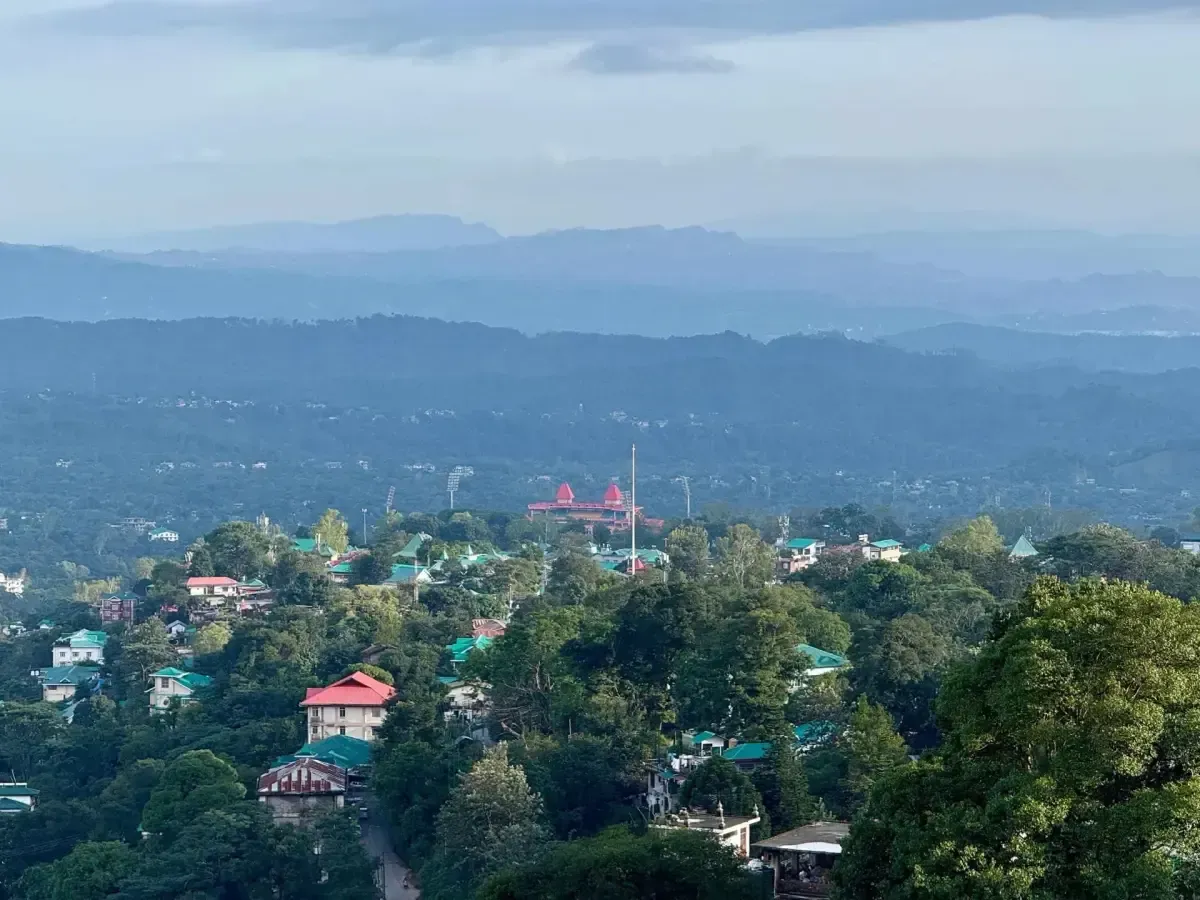 A hill town spread across a wooded slope below, houses with red and blue roofs among the trees, ridges fading into haze behind