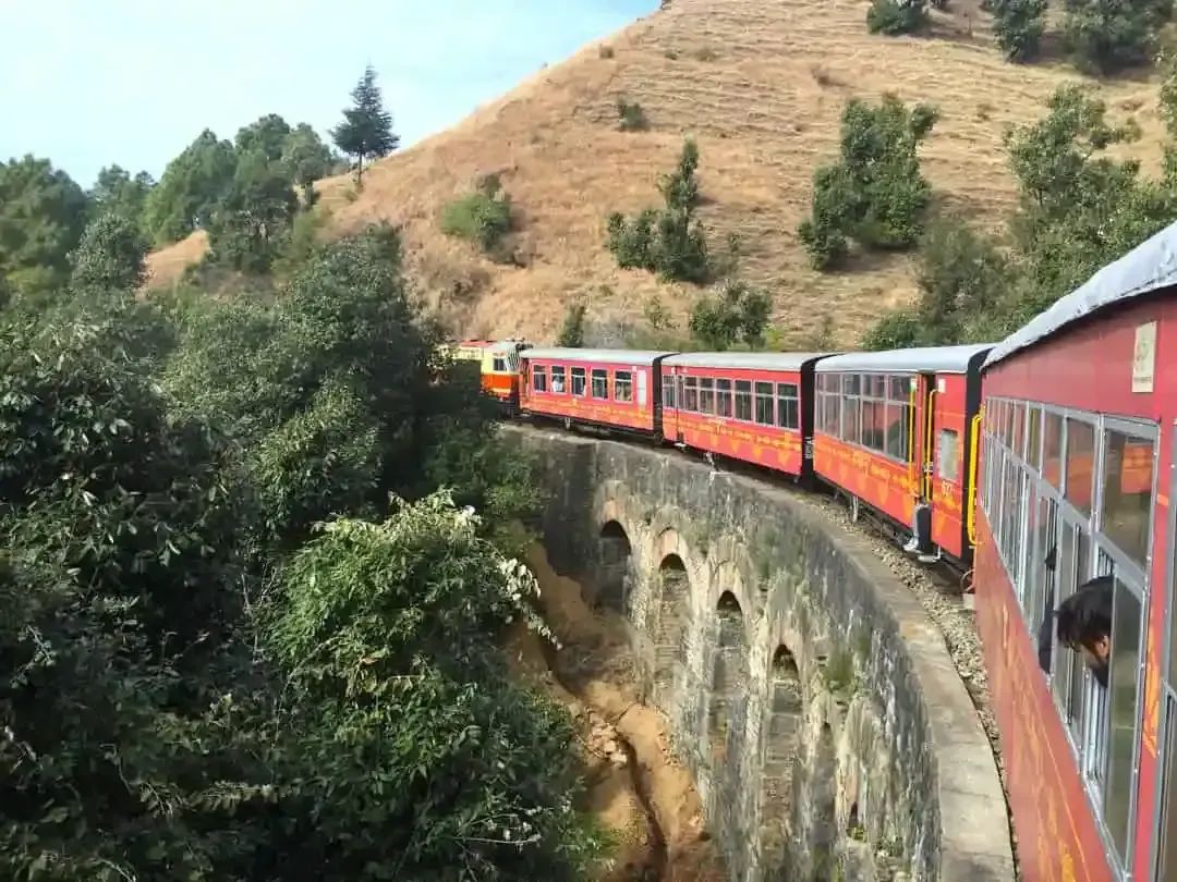 A narrow-gauge train in red livery curving across a multi-arched stone viaduct on a wooded hillside, photographed from a carriage window further back along the same curve.