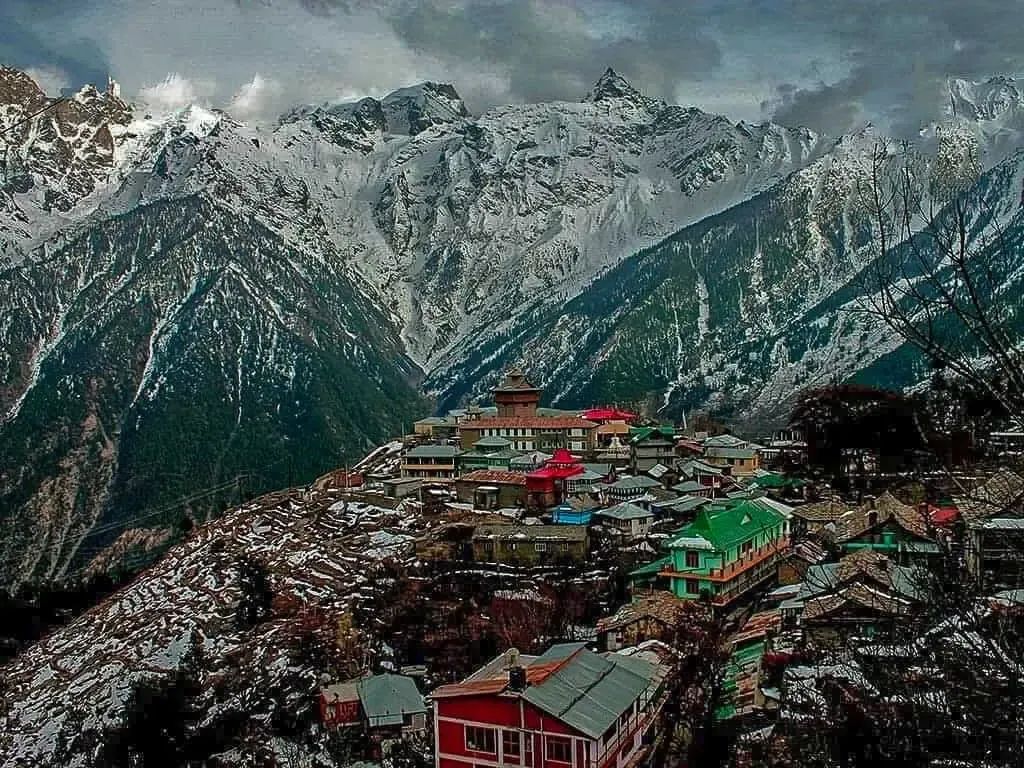 A village of tin roofs and a slate-roofed temple stacked on a wooded spur, a wall of snow peaks filling the sky behind it.