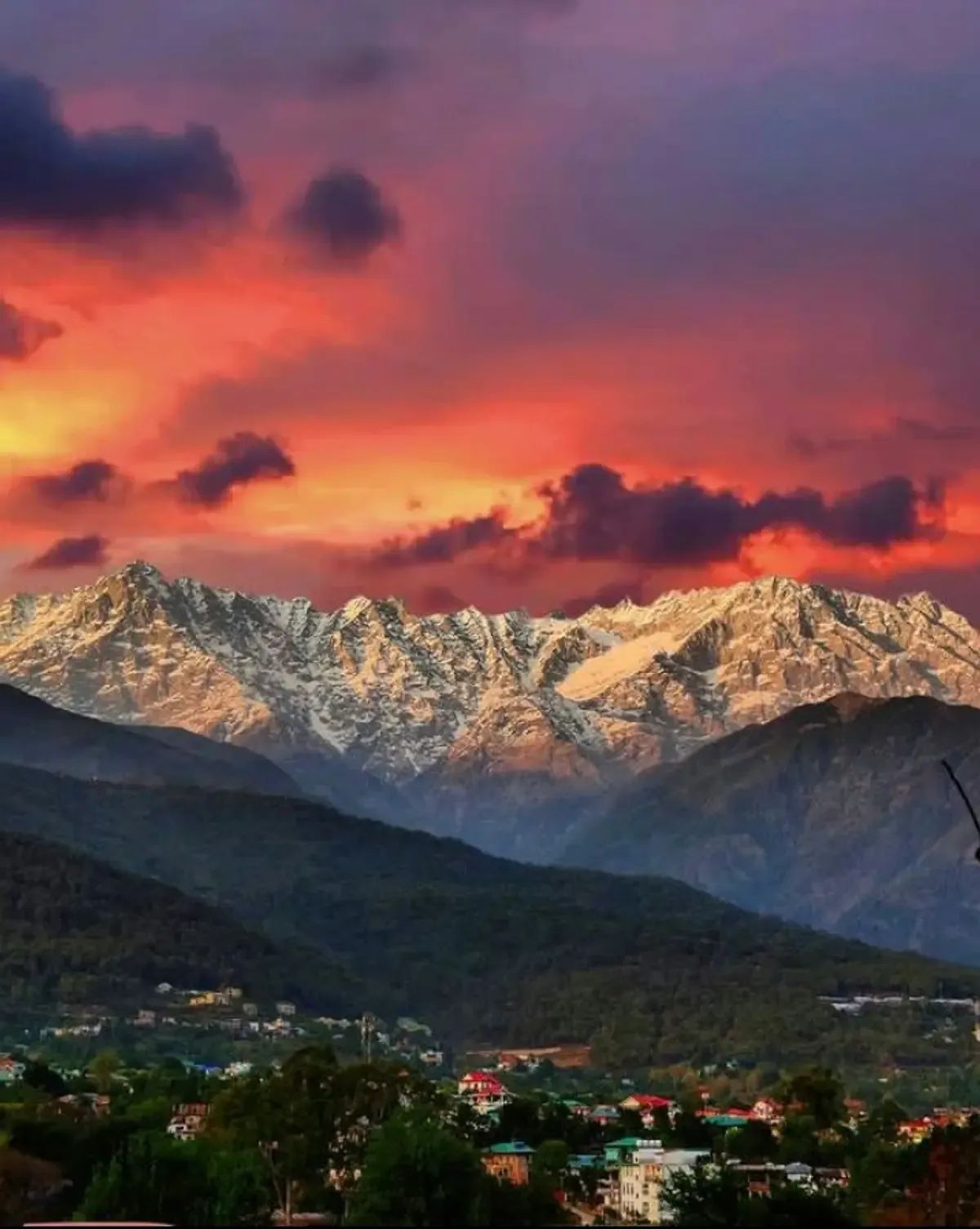 A long wall of snow-covered peaks catching low red light, with forested ridges and the roofs of a town below, under a sky of orange and dark cloud.