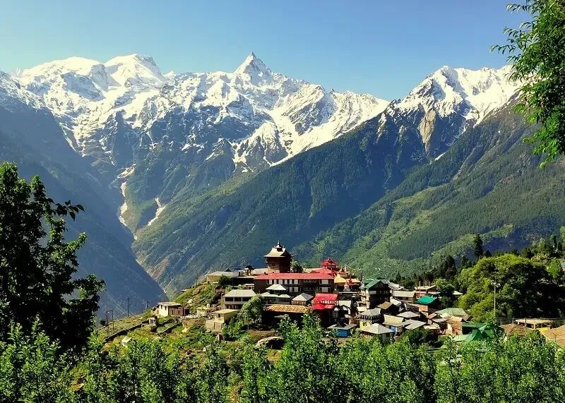 A village of slate-roofed houses stacked on a green spur above a wooded valley, with a long wall of snow peaks catching the light directly behind it.