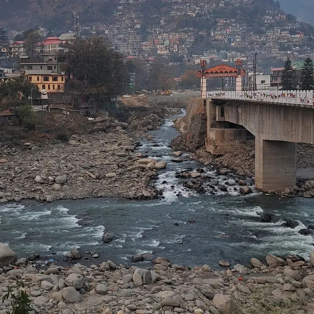 A shallow river running fast over a wide bed of pale boulders, with a concrete road bridge crossing it and the town stacked up the bank beyond.