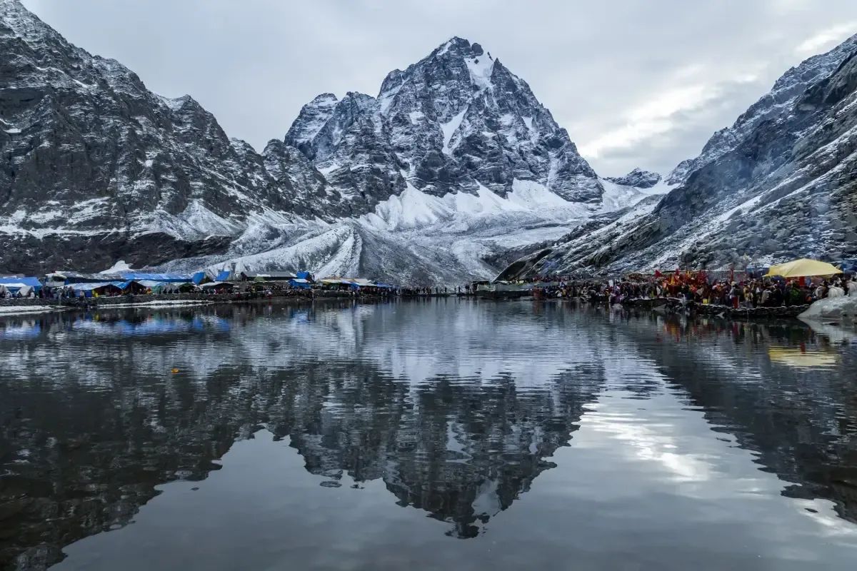 A dark snow-streaked peak standing above a still lake, with lines of tarpaulin shelters along the far shore and the whole scene reflected in the water.