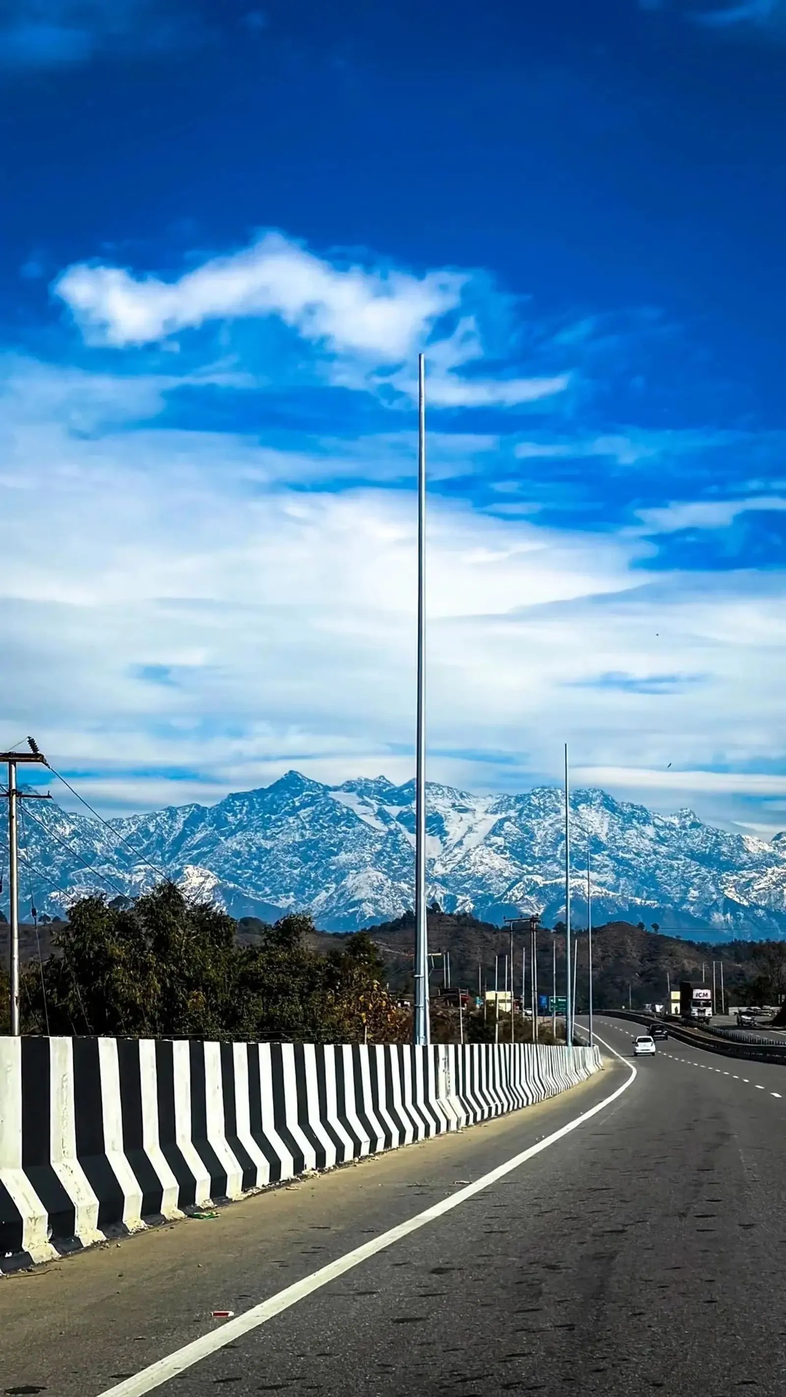A divided highway curving away past a black-and-white striped concrete barrier and a row of tall lamp posts, with a long wall of snow-covered peaks filling the horizon under deep blue sky.