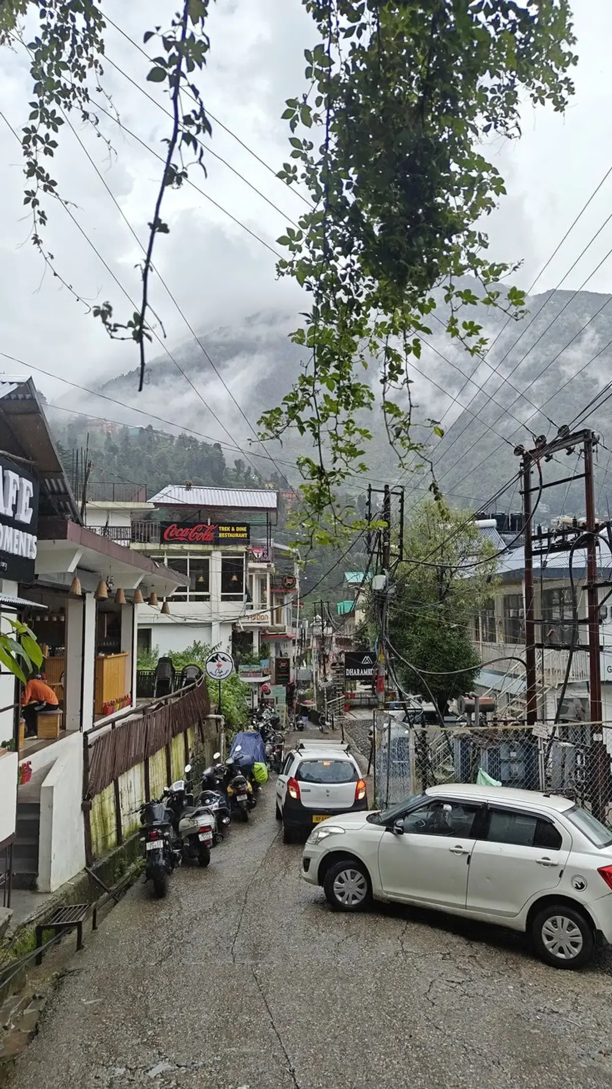 Shopfronts and a restaurant sign packed along a slope below a hillside lost in cloud, power cables strung in every direction across the frame