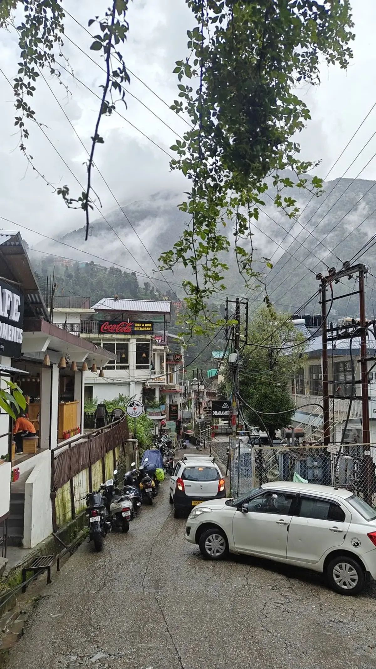 A McLeod Ganj street in low cloud, shopfronts and prayer flags on both sides and the road dropping away into mist.