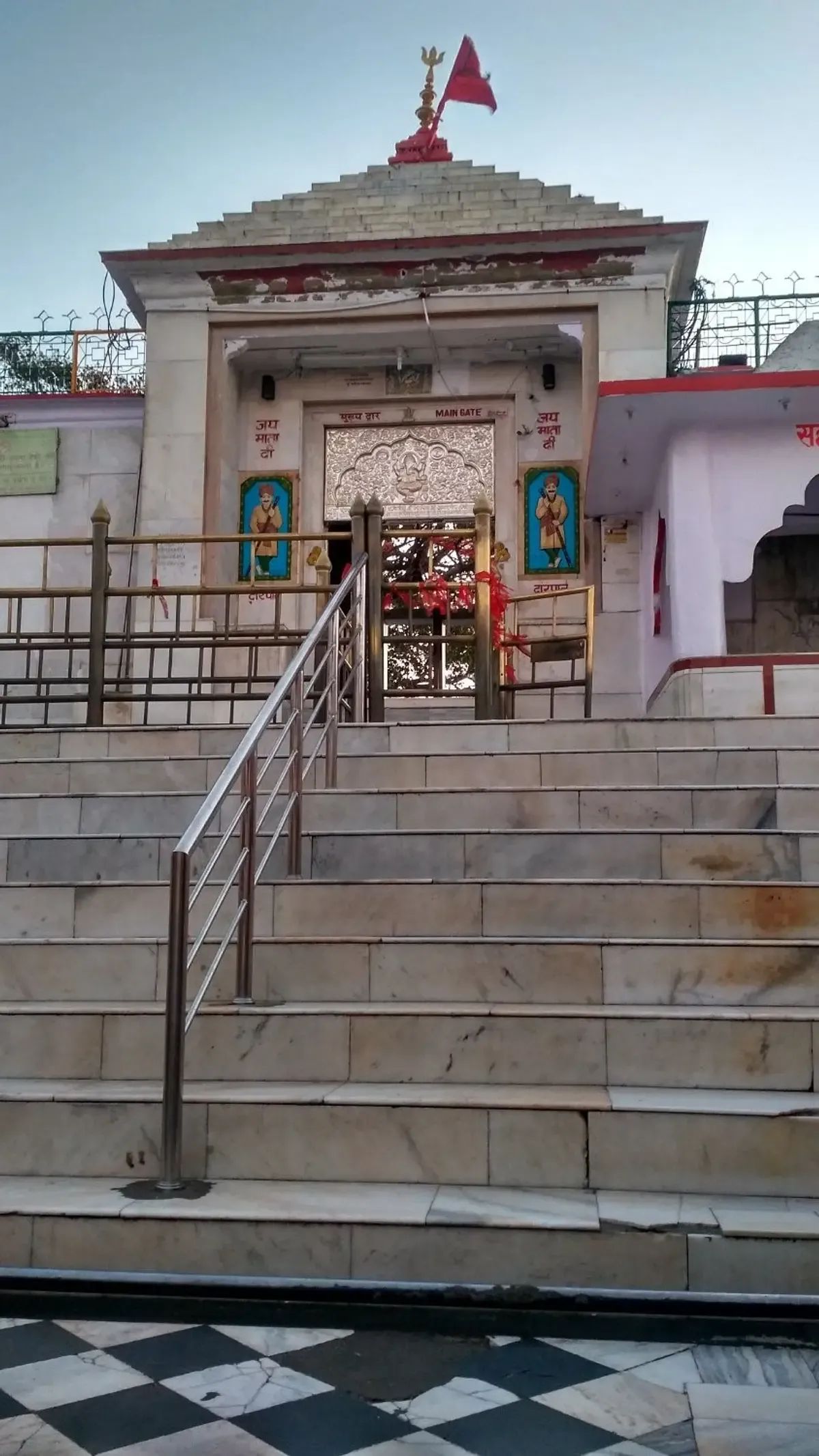 A temple gateway of pale stone with a stepped roof, a red pennant and trident at its peak, painted guardian figures either side of a silver-embossed doorway, reached by a broad flight of marble steps with a steel handrail.