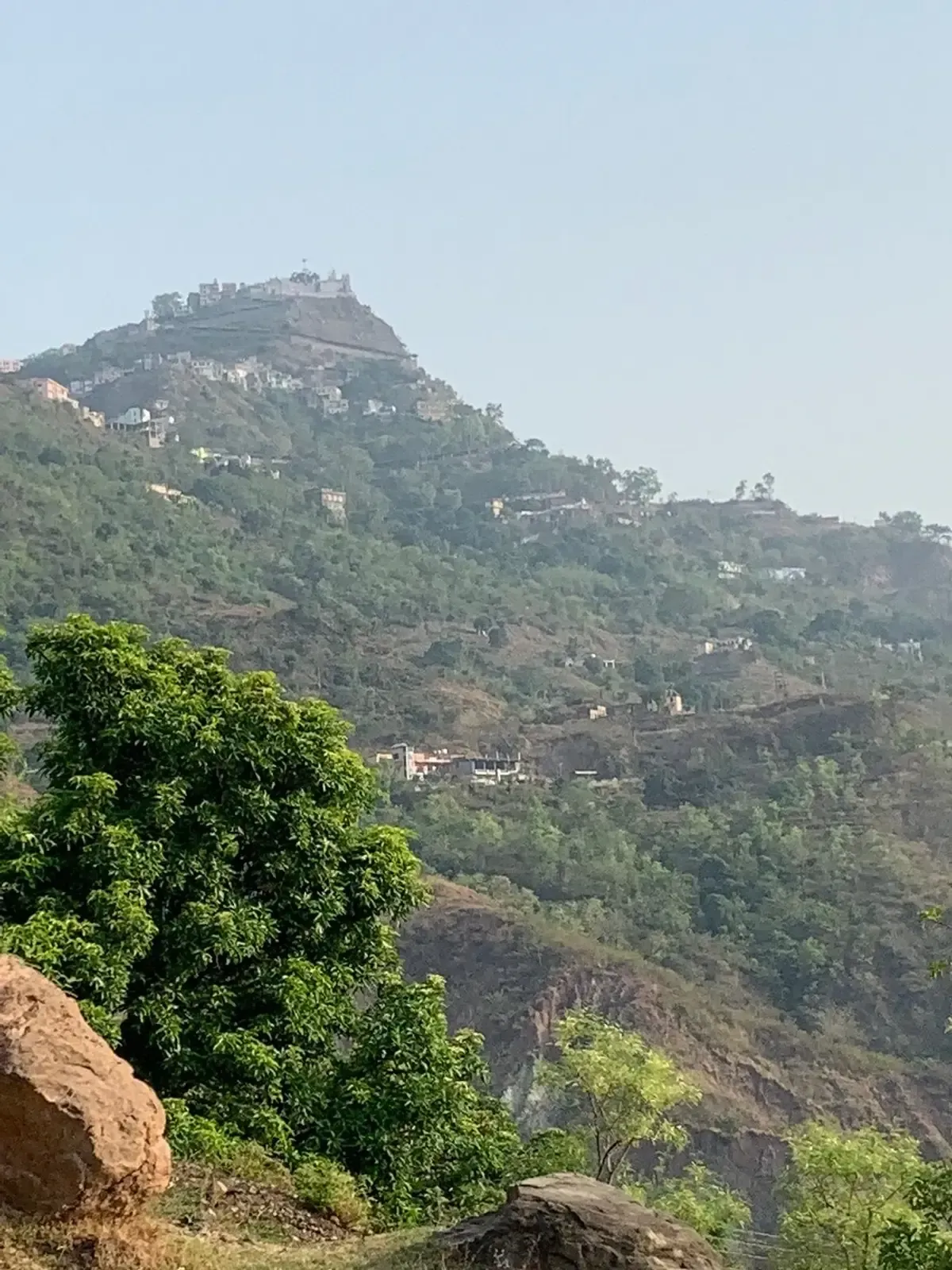 A temple complex of white and ochre buildings along the top of a steep wooded hill, houses scattered on the slopes below