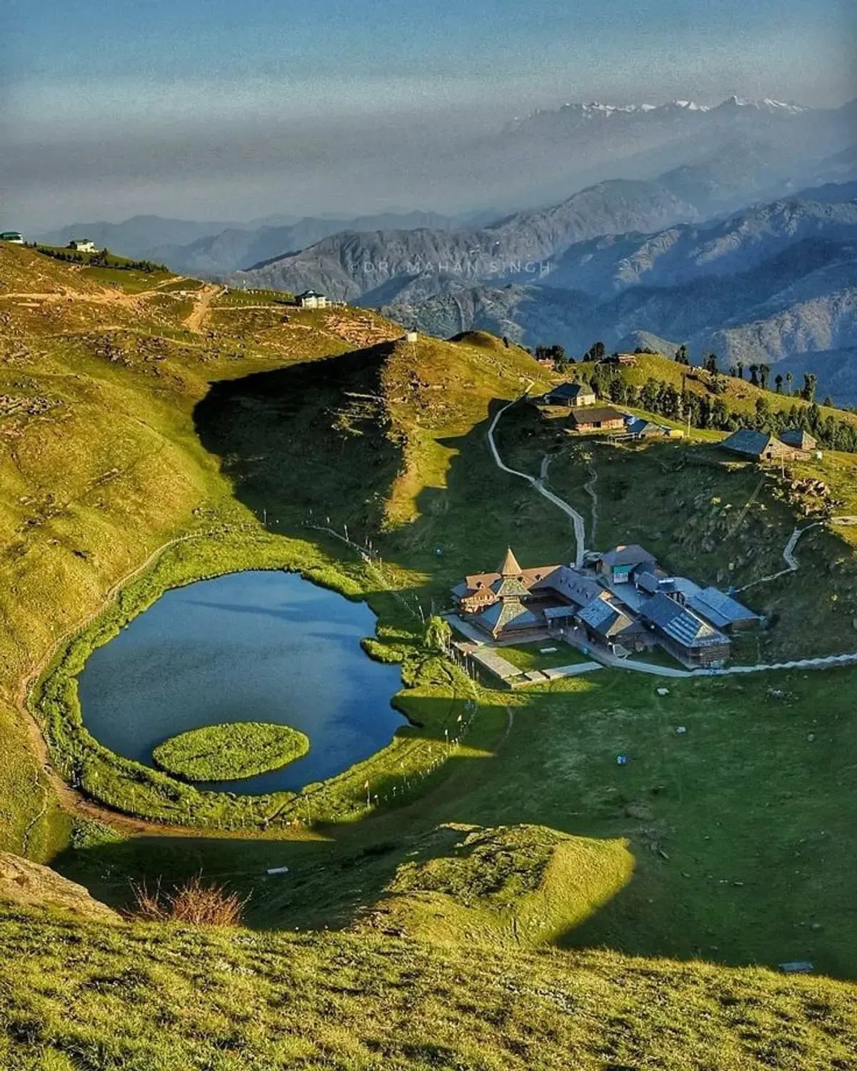 A small round lake with a green floating island on it, held in a bowl of meadow, with a tiered wooden temple on the slope beside the water.