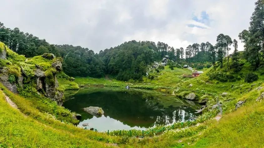 A small dark lake sitting in a bowl of cropped grass, ringed by boulders and old forest, with a low building among the trees on the far bank.