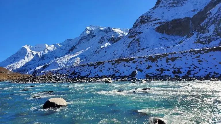 A wide braided river running over pale boulders in bright sun, with a long snow-covered ridge standing directly behind it under a clear sky.