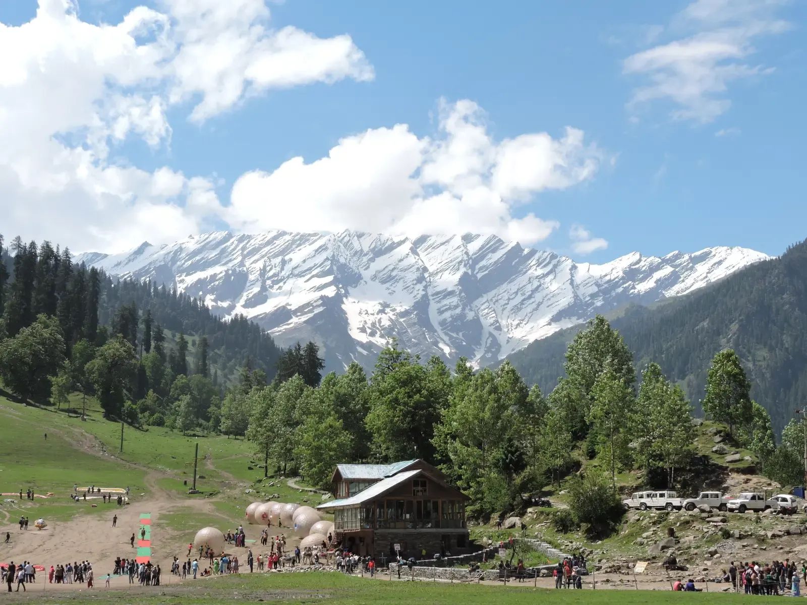 A broad grass meadow with small figures spread across it, a timber building and parked vehicles along its edge, conifers on the slope beyond and a wall of snow closing the head of the valley.
