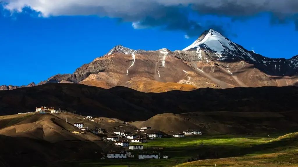 Whitewashed flat-roofed houses and green barley strips of a high village, spread along a bare brown slope under a snow-capped peak.