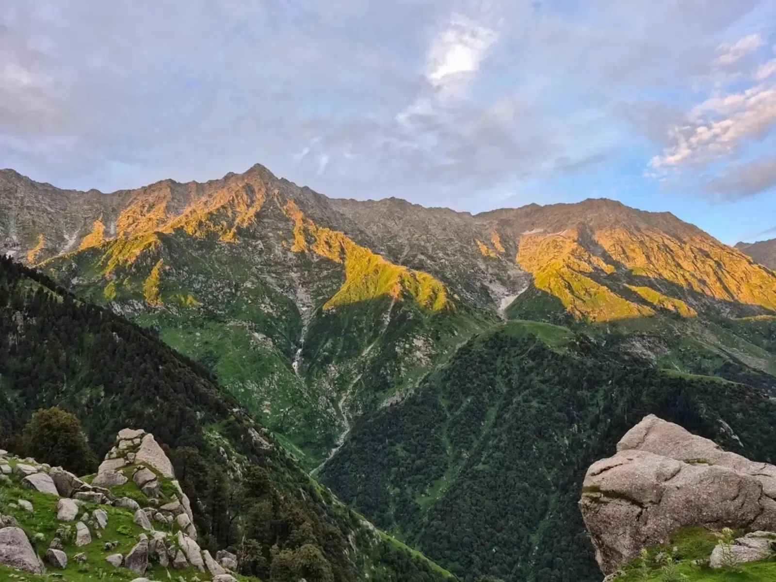 The last sun on a high ridge, the grass and rock along the top of it lit orange while the wooded valley below has gone into shadow, with boulders in the foreground.